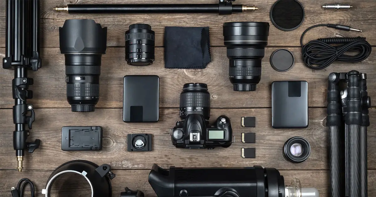 Equipamento de vídeo sobre uma mesa de madeira: câmera, lentes, tripés, baterias, cartões de memória e cabos