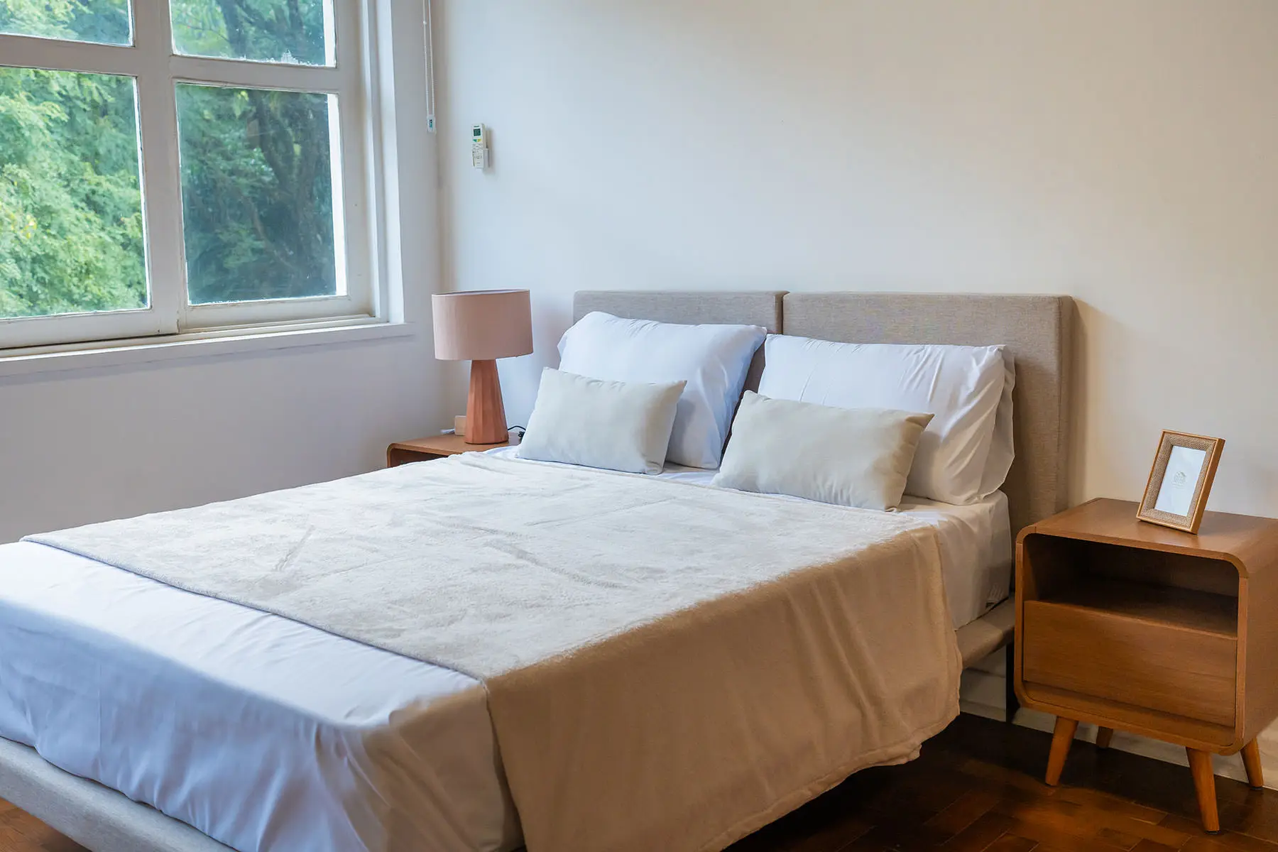 Ambiente quarto do estúdio: cama de casal arrumada com cabeceira estofada, abajur no criado-mudo e janela com vista para árvores