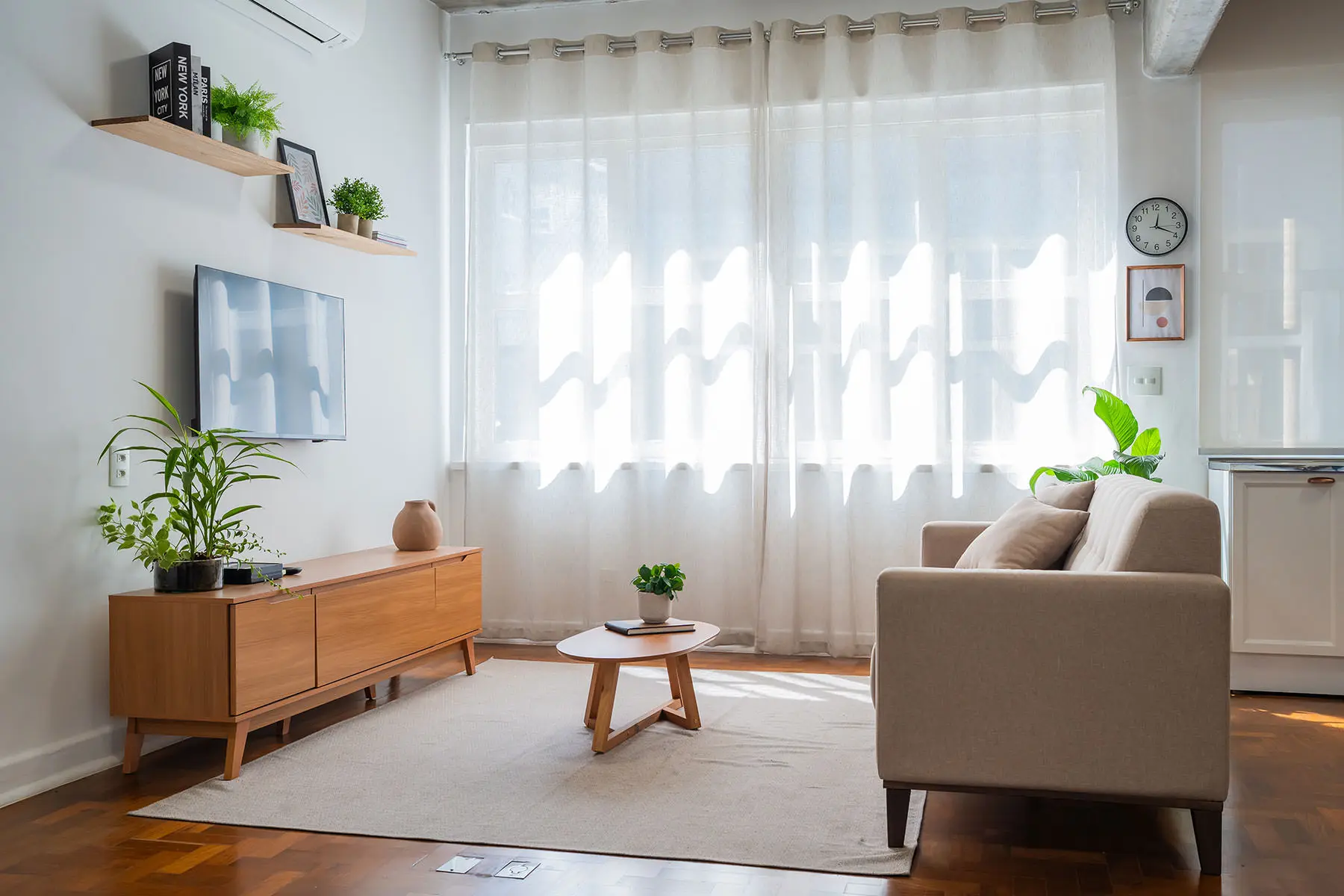 Ambiente sala do estúdio: sofá bege, tapete claro, mesa de centro, TV na parede e janela com cortina de luz filtrada